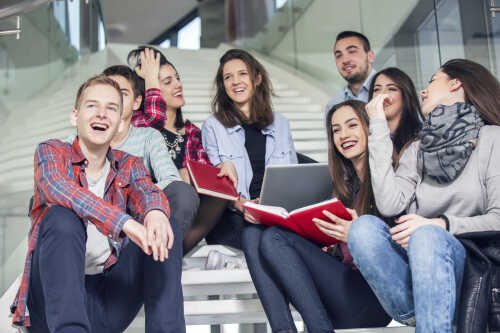 Several students are sitting together on a staircase, they laugh, talk and point