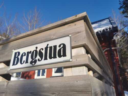 Picture of the sign hanging on the fence. The sign it says "Bergstua", we see the red cabin in the b