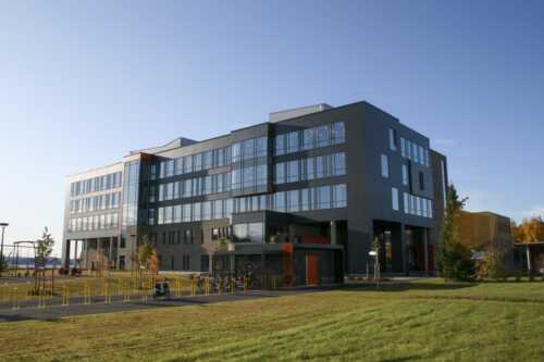 Stort grått bygg, omtrent 4 etasjer. Det er mange vinduer rundt hele bygget. Gresset er grønt, og 