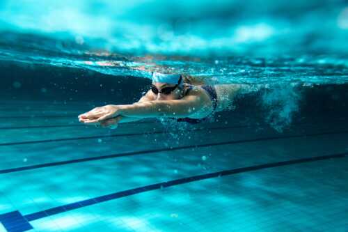 Kvinne med dykkebriller og badehette svømmer under vann. 