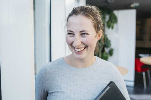Bilde av en smilende, kvinnelig student i grå genser. Hun holder en bok i hånden.