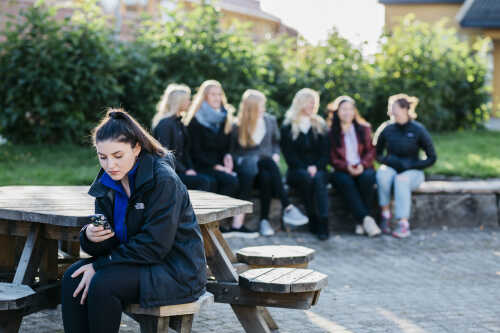 Kvinnelig student sitter utendørs og ser på mobilen. En gjeng med kvinnelige studenter i bakgrunne