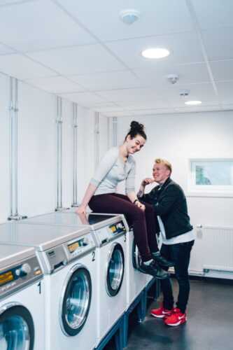 Woman sitting on the washing machine and man leaning on it, shows the laundry